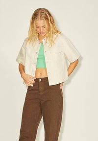 Casaco creme cropped com mangas curtas, dois bolsos no peito, sobre um top cropped verde e calças castanhas de cintura alta. Textura suave.