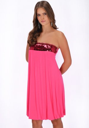 Jonge vrouw met lang bruin haar, gekleed in een strapless roze jurk met een rood geborduurde top, staand met haar handen achter haar rug tegen een witte achtergrond.