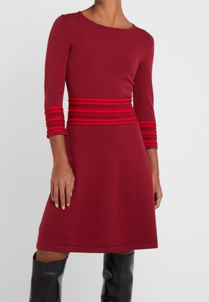 Femme portant une robe bordeaux longueur genou avec des manches trois-quarts et des rayures horizontales rouges à la taille et aux poignets.