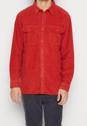 Homme portant une chemise en velours côtelé rouge à manches longues avec deux poches poitrine, associée à un pantalon gris foncé, debout devant un fond blanc.