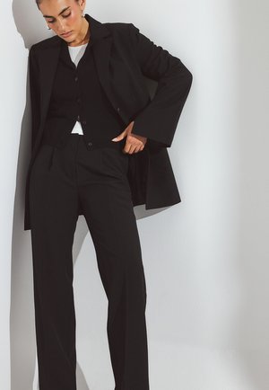 Schwarze maßgeschneiderte Hosen kombiniert mit einem passenden langen Mantel und einem schwarzen gefütterten Weste. Der Look zeichnet sich durch klare Linien und eine strukturierte Silhouette aus.