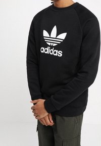 Svart sweatshirt med rund hals, ribbade ärmslut och nederkant, med en stor vit Adidas-logotyp och tre ränder emblem på framsidan.