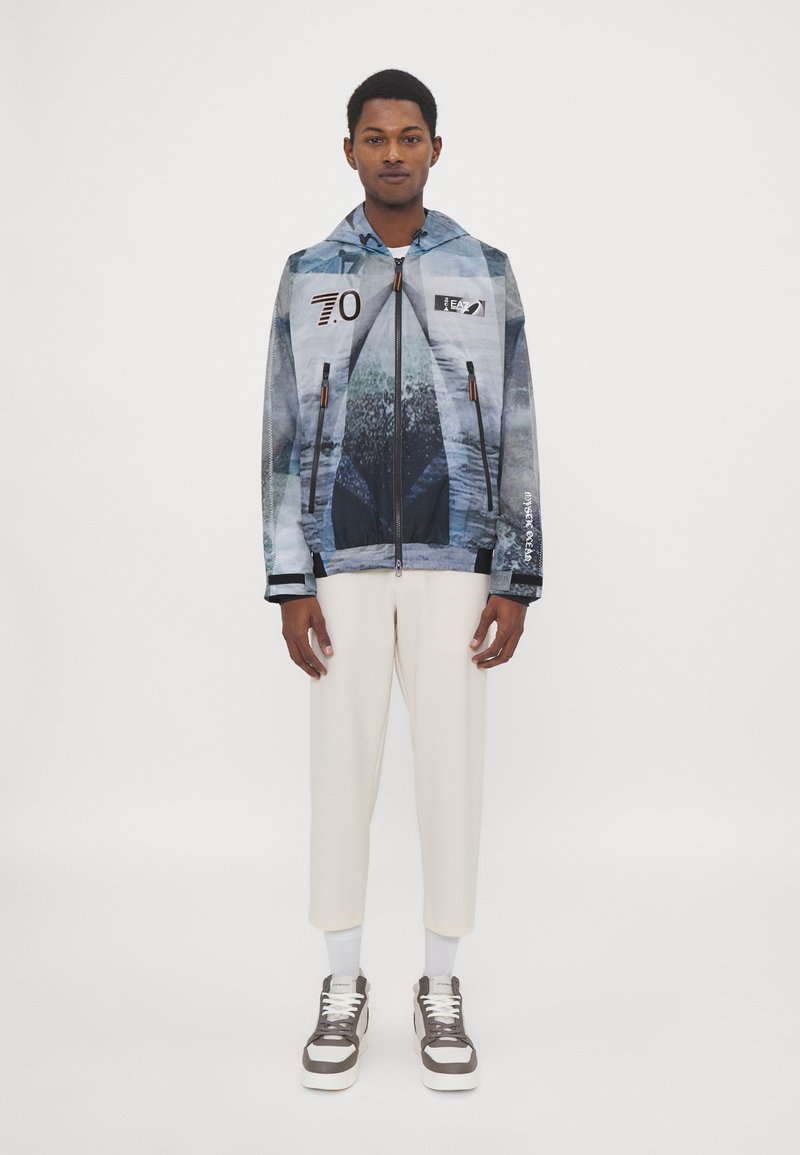 Blaue und graue Jacke mit abstraktem Muster, Kapuze, Reißverschlüssen und Logodetails, kombiniert mit weißen tapered Hosen und grauen Sneakern.