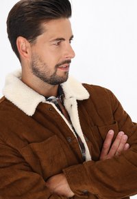 Braune Cord-Jacke mit einem weißen Schaffellkragen, versehen mit einer knöpfbaren Vorderseite, zwei Brusttaschen und einem strukturierten Stoff.