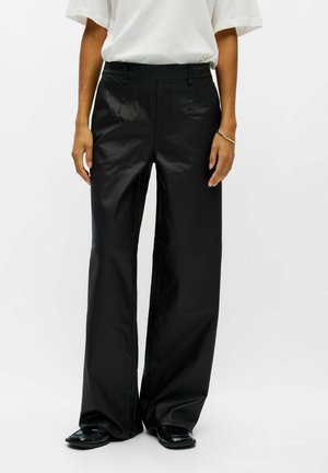 Modèle portant un pantalon noir à jambes larges, un T-shirt blanc et des chaussures noires sur un fond blanc uni.