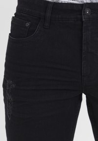 Mustad teksapüksid, millel on kitsas lõige, vasaku reie õrn kulutatus ja klassikaline viie taskuga disain metallist detailidega.