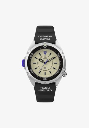 Ura Timex INDIGLO s kremnim številčnikom, črnimi silikonskimi trakovi in srebrnim ohišjem iz nerjavečega jekla. Ima svetleče kazalce in vrteči se bezel.
