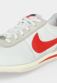 Λευκό αθλητικό παπούτσι Nike με κόκκινο λογότυπο Swoosh, λευκά κορδόνια και μπεζ σουέντ περιθώριο, εμφανίζεται σε κοντινή λήψη σε ανοιχτό φόντο.