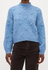 Jersey de punto azul claro con cuello alto, hombros caídos y patrones de estrellas texturizados. Con dobladillo y puños de canalé. Combinado con pantalones de pana marrón oscuro.