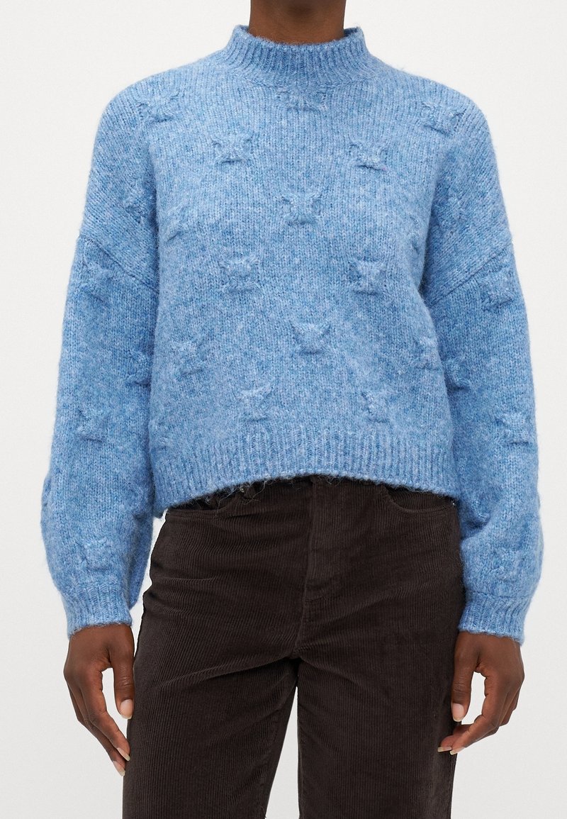 Jersey de punto azul claro con cuello alto, hombros caídos y patrones de estrellas texturizados. Con dobladillo y puños de canalé. Combinado con pantalones de pana marrón oscuro.