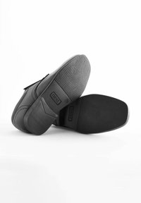 Chaussures en cuir noir avec une finition lisse, bande velcro, semelles en caoutchouc texturé et logo de la marque en relief sur la semelle.