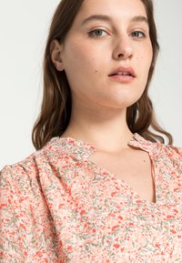Blusa floral com um padrão em coral e creme, com um decote franzido e um tecido leve que cai suavemente sobre o ombro.