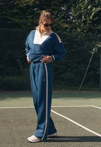 Blauer Sportanzug mit weißen Akzenten; Reißverschlusskragen, lockere Passform und gerippte Bündchen. Getragen mit weißen Sneakers auf einem Sportplatz.