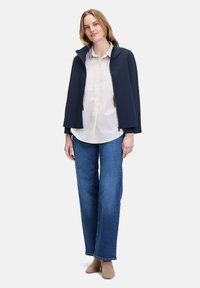 Marineblaues Cape mit Kapuze, getragen über einer weißen Bluse mit Streifen, kombiniert mit blauen weiten Jeans und beigen Schuhen. Glatter Stoff, minimalistisches Design.