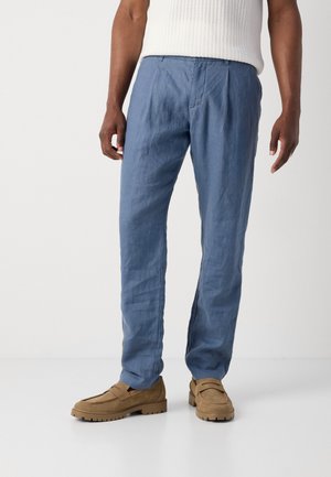 Lichtblauwe linnen broek met een relaxte fit, voorzien van plooien aan de voorkant en zijzakken, gecombineerd met bruine suède instappers en een geribde witte top.