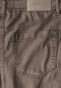 Los pantalones de mezclilla marrones presentan un bolsillo trasero con una costura sutil y una etiqueta rectangular marcada "STREET ONE - DENIM ATELIER."