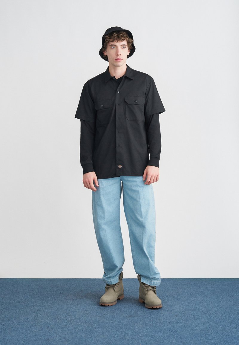 Chemise noire à manches courtes avec deux poches poitrine, jean ample bleu clair, bottines grises et chapeau seau noir. Texture de tissu lisse.