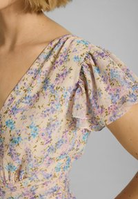 Frau in einem pastellfarbigen Blumenkleid mit gerafften kurzen Ärmeln und V-Ausschnitt vor einem schlichten grauen Hintergrund.