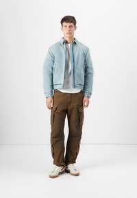 Veste bomber en denim bleu clair, t-shirt gris et pantalon cargo marron avec poches. Des baskets blanches avec des accents marron complètent la tenue.
