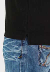 Fekete pamut póló oldalsó hasítékokkal, világoskék farmernadrággal, amely elhasználódott mintákkal és kontrasztos narancssárga varrási részletekkel rendelkezik.