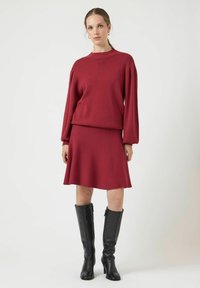 Czerwona dzianinowa sweter z bufkami na długich rękawach, w połączeniu z rozkloszowaną czerwono dzianinową spódnicą. Kobieta nosi czarne botki za kolano. Czyste tło.