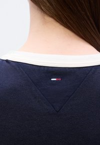 Μπλε σκούρο φούτερ με υφή ύφανσης, που διαθέτει έναν αντίθετο λευκό γιακά και ένα μικρό κόκκινο, λευκό και μπλε λογότυπο ραμμένο στο πίσω μέρος.