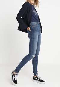 Modré skinny džíny s vysokým pasem a roztrženými detaily, zkombinované s námořnickým tričkem a černým blazerem, a černobílými teniskami.