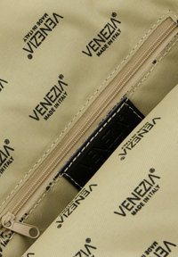 Beige stoftas indvendig med gentagne "VENEZIA" logoer i sort, en glat lynlås og et sort brandmærke. Tekstureret overflade.