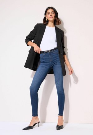 Kvinde iført en sort blazer, hvid top, blå skinny jeans med sort bælte og sorte spidse hæle står mod en ensfarvet væg.