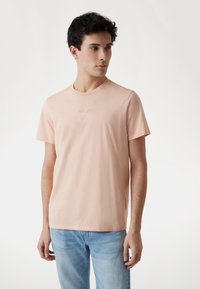 Világos rózsaszín pamut póló, kerek nyakkivágással, a mellrészen finoman dombornyomott "GUESS" felirattal, világoskék farmernadrággal párosítva.