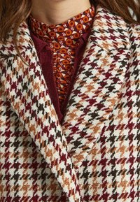 Wool houndstooth-patterned coat with burgundy, orange, and white colors; features a broad collar and textured fabric.