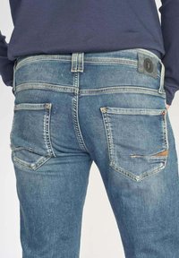 Les jeans en denim bleu présentent un design ajusté, des coutures décoratives, deux poches arrière avec des accents et une étiquette en cuir sur la ceinture.
