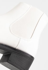 Bottine blanche à motif crocodile texturé, avec un panneau élastique et un petit talon noir. Surface lisse et légèrement brillante.