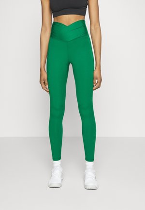 Femme portant des leggings taille haute vert vif, des baskets blanches et un crop top noir, debout devant un fond blanc uni.