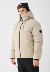 Jachetă puffer bej cu guler înalt, fermoar negru și �șnururi ajustabile. Are o suprafață texturată și un accent cu logo pe mânecă.