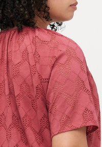 Top color rosa con un motivo in pizzo testurizzato caratterizzato da forme geometriche e accenti floreali. Maniche corte e scollatura curva.