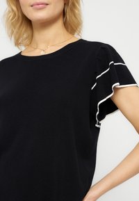 Γυναίκα με μαύρο κοντομάνικο τοπ με άσπρη επένδυση στα φουσκωτά μανίκια και χρυσό κρεμαστό κολιέ, μπροστά σε απλό ανοιχτό φόντο.