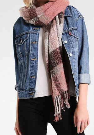 Femme portant une veste en denim bleu, une chemise blanche, un pantalon noir et une épaisse écharpe à carreaux roses et bruns avec des franges.