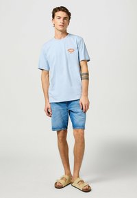 Ljusblå bomulls T-shirt med kort ärm och en grafisk bild framtill. Kombinerad med denimshorts och beige sandaler med en rosettdetalj.