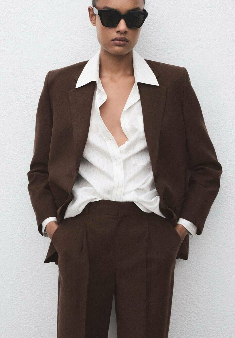 Bruin op maat gemaakt pak met een enkelgeknopte blazer en rechte broek. Witte shirt met verticale strepen eronder. Zwarte zonnebril.