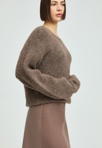 Frau mit glattem, dunkelbraunem Haar, die einen flauschigen braunen Pullover und einen passenden braunen Rock trägt, steht vor einem schlichten, hellen Hintergrund.
