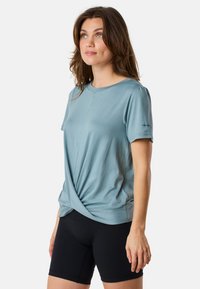 Ljusblå topp med kort ärm och ett tvinnat framsidesdesign samt rund halsringning, i kombination med svarta shorts. Tillverkad av ett mjukt, elastiskt tyg.