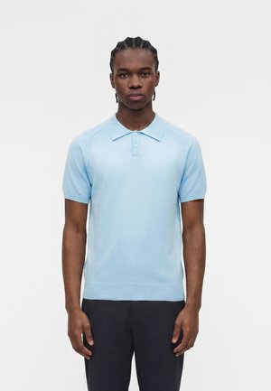 Homme aux cheveux tressés portant un polo bleu clair à manches courtes et un pantalon foncé, debout sur un fond blanc uni.