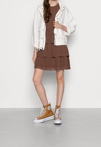 Weiße Jacke über einem braun karierten, gestuften Kleid, kombiniert mit tanfarbenen High-Top-Sneakers mit weißen Sohlen und Schnürsenkeln. Glatte, lässige Texturen.