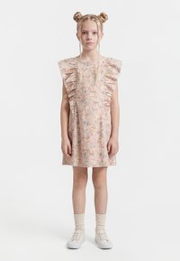 Jong meisje met blond haar in twee knotjes en vlechtjes, gekleed in een korte bloemenjurk met ruches, crèmekleurige sokken en witte sneakers, staand in een rechte houding.