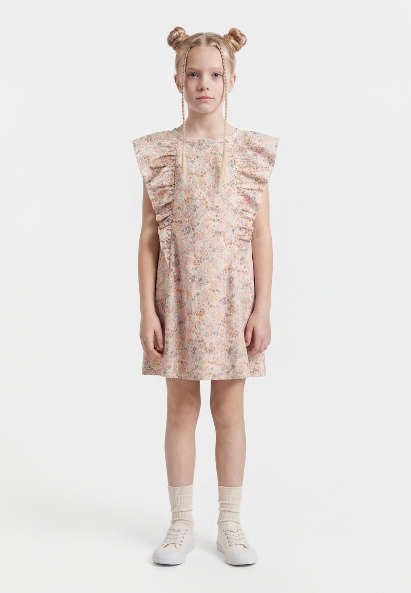 Jong meisje met blond haar in twee knotjes en vlechtjes, gekleed in een korte bloemenjurk met ruches, crèmekleurige sokken en witte sneakers, staand in een rechte houding.