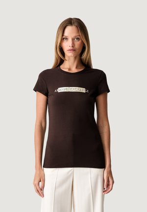 Mujer de cabello largo lleva una camiseta negra ajustada Armani Exchange y pantalones blancos, de pie frente a un fondo claro y sencillo.