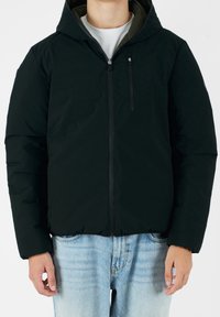 Sort hættepufjakke med frontlynlås, to sidelommer og en glat tekstur. Båret over en lys grå skjorte og blå denim.