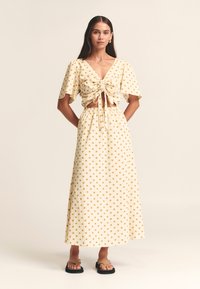 Rumena kompletna obleka z belimi pikami, ki vključuje kratki zgornji del z zavezovanjem spredaj in kratkimi rokavi ter usklajeno dolgo krilo, izdelano iz lahkega materiala.