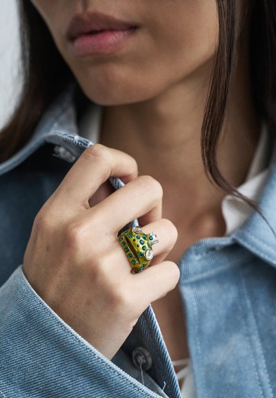 Yellow ring with green and blue gemstones, featuring a textured surface. Worn on a hand grasping a denim jacket collar.